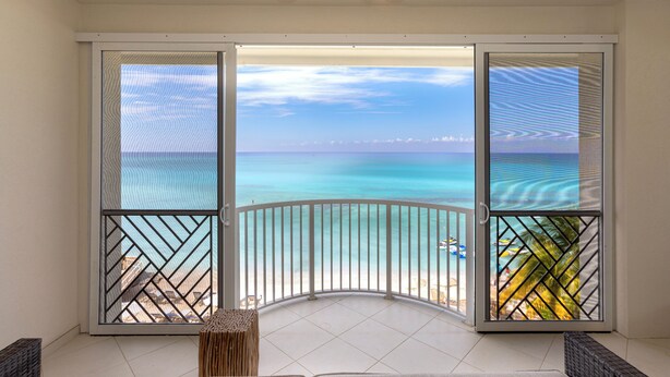 A balcony with a view of the ocean, a cushioned seating area, and a small table. - Beachfront 4BR/4BA | Sleeps 10, Ocean Views, Pool (Seven Mile Beach)