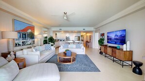 A spacious living room with a large flat-screen TV, a sofa set, a coffee table, and a dining area with a wooden cabinet. - Beachfront 4BR/4BA | Sleeps 10, Ocean Views, Pool (Seven Mile Beach)