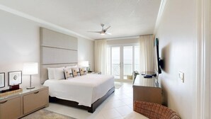 A modern hotel room with a large bed, a flat-screen TV, a ceiling fan, and a balcony with curtains. - Beachfront 4BR/4BA | Sleeps 10, Ocean Views, Pool (Seven Mile Beach)