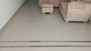 A spacious living room with a beige sectional sofa and ottoman, white walls, and a tiled floor.