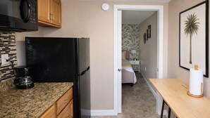A compact kitchen with a refrigerator, microwave, and coffee maker. A bedroom with a bed and nightstand is visible through an open door. - Guest Suite: Close to Beach & Downtown St. Pete (Pinellas Park)