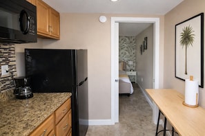 A compact kitchen with a refrigerator, microwave, and coffee maker. A bedroom with a bed and nightstand is visible through an open door.