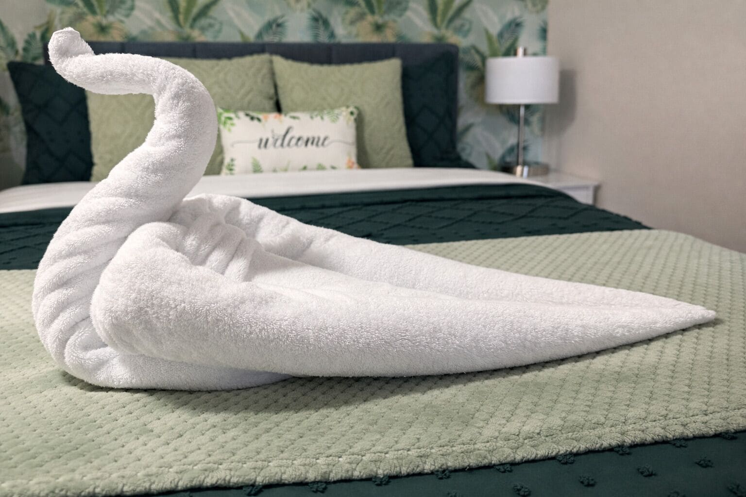 Una toalla con forma de cisne sobre una cama con una colcha verde y una almohada con el texto 'welcome'.