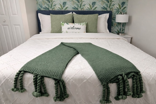 A bed with a white quilt, a green knitted throw, and a 'welcome' pillow.