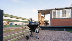 A barbecue grill on a gravel patio with a wooden fence and a building in the background. - Romantic Houseboat with out of this World Star Gazing (Alpine)