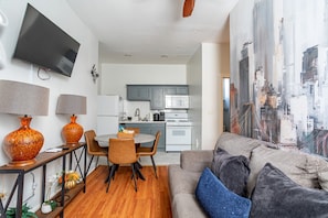 A modern living room with a flat-screen TV, a dining area with a table and chairs, a kitchen with grey cabinets, and a large cityscape painting on the wall.