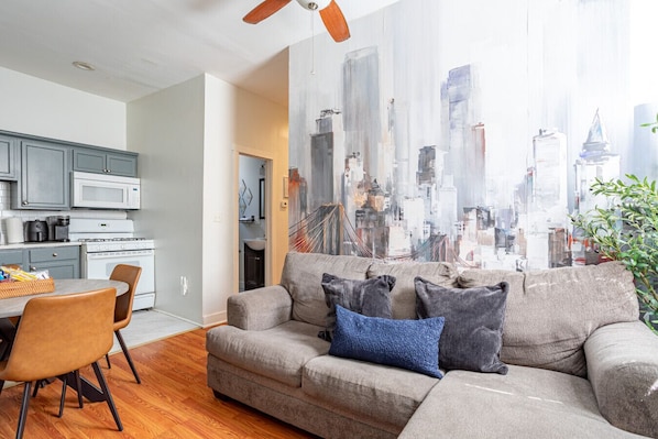 A modern living room with a gray sofa, a dining table with chairs, a kitchen area, and a cityscape view.