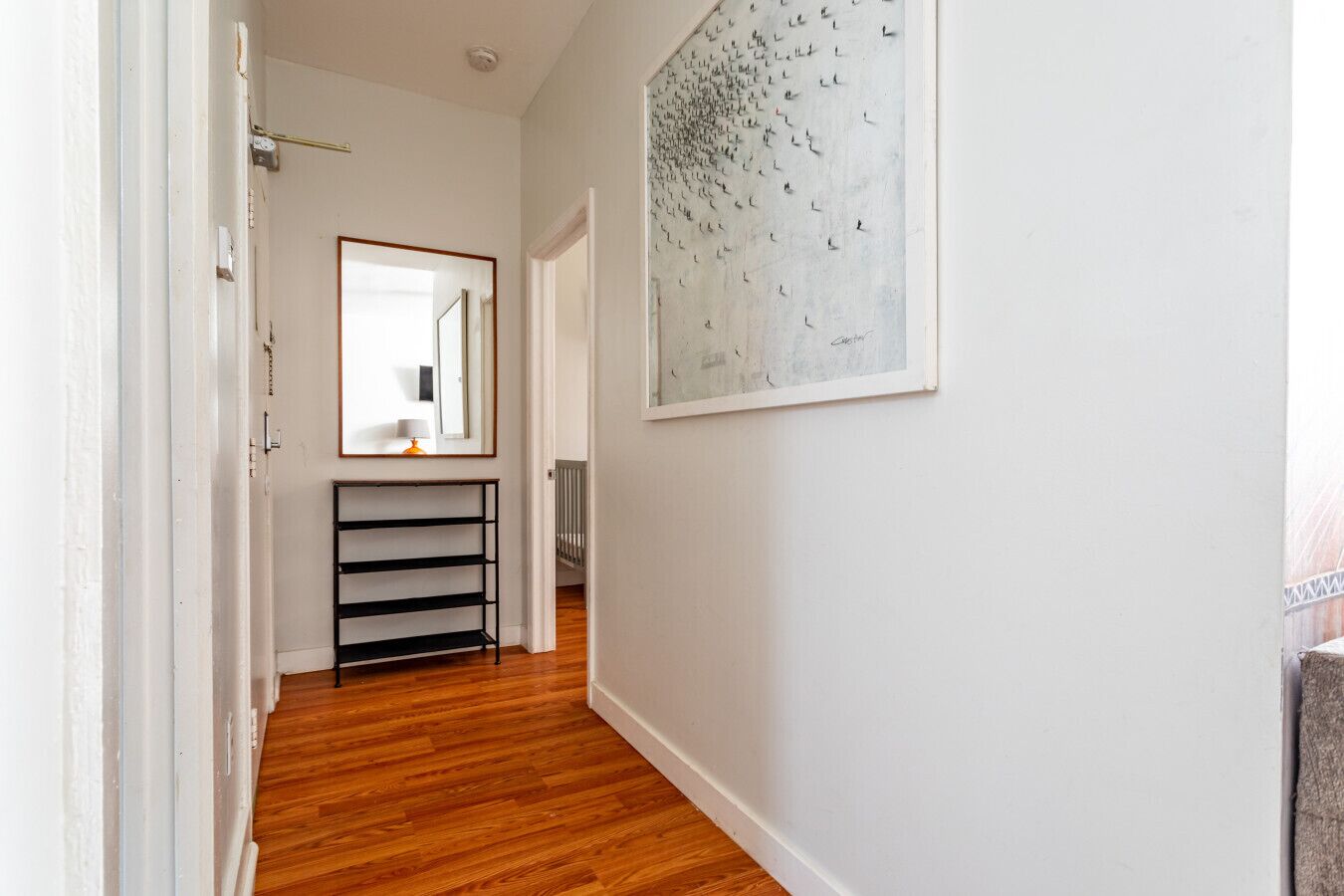 A hallway with wooden flooring, a black metal shelf, a large abstract painting, and a white door.