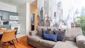 A modern living room with a gray sofa, a dining table with chairs, a kitchen area, and a cityscape view.
