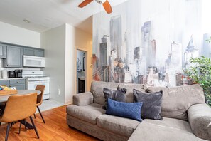 A modern living room with a gray sofa, a dining table with chairs, a kitchen area, and a cityscape view.