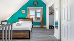 A bedroom with a sloped ceiling, a bed, a window with curtains, and a view of buildings outside.