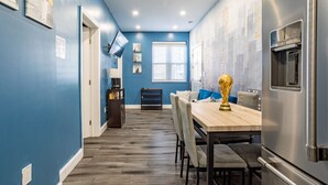 A narrow hallway with a dining area featuring a wooden table and chairs, a TV mounted on the wall, and a refrigerator on the right.