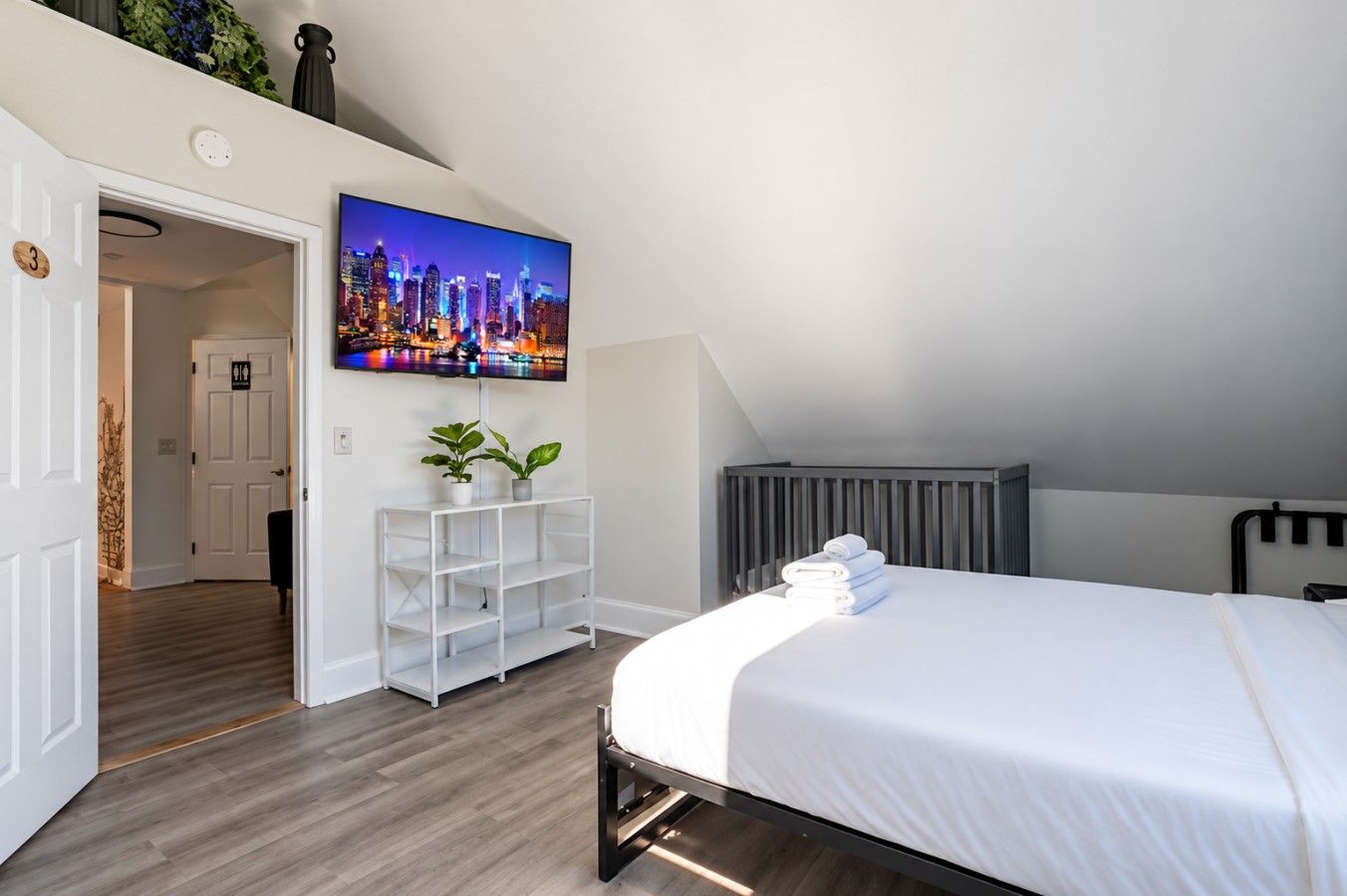 Ein modernes Schlafzimmer mit einem Bett, einem an der Wand befestigten Fernseher, einem weißen Konsolentisch mit Pflanzen und einer Heizung.