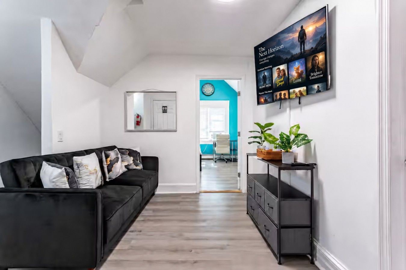 A modern living room with a black sofa, a coffee table with plants, a TV mounted on the wall displaying movie posters, and a hallway leading to another room.