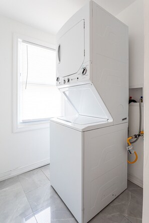 A compact, white washing machine in a room with a window and tiled floor.