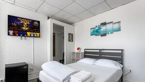 Ein Hotelzimmer mit einem Bett, einem kleinen Kühlschrank, einem Fernseher, der das Poster der FIFA Fussball-Weltmeisterschaft 2026 zeigt, und einer Wand mit drei gerahmten Kunstwerken.