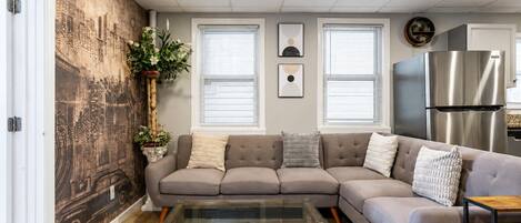 A living room with a sectional sofa, a glass coffee table, a cityscape mural, and a stainless steel refrigerator.