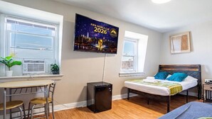 Une chambre avec un lit, un bureau avec des chaises, un téléviseur fixé au mur et une petite enceinte noire.