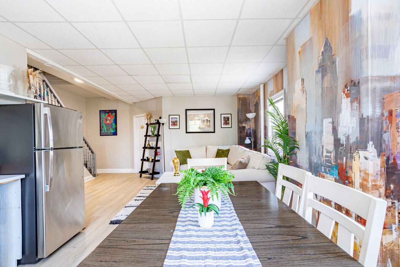 A modern kitchen with a stainless steel refrigerator, a dining area with a wooden table and chairs, a white sofa, and a cityscape mural on the wall.