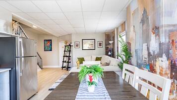 A modern kitchen with a stainless steel refrigerator, a dining area with a wooden table and chairs, a white sofa, and a cityscape mural on the wall.