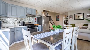 Une cuisine moderne avec des meubles gris, un réfrigérateur en acier inoxydable et un coin repas comprenant une table en bois et des chaises blanches.