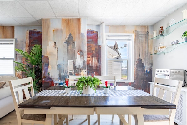 A dining area with a wooden table, white chairs, potted plants, and a cityscape mural on the wall.