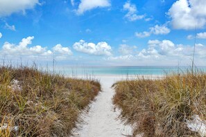 Un sentier bordant la plage, menant à l’océan, avec un ciel dégagé et quelques nuages.