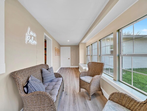 Une pièce spacieuse, meublée de mobilier en osier, dotée de grandes fenêtres et ornée d’un panneau lumineux au néon sur lequel on peut lire « One Palm ».