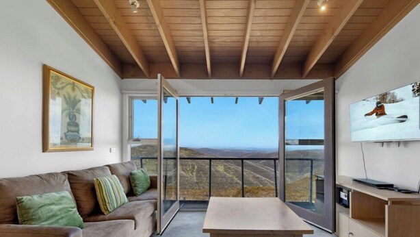 A modern living room with a wooden ceiling, a sofa, a table, and a large window offering a scenic view. - White Crystal 209- ski-in ski-out (Mount Hotham)