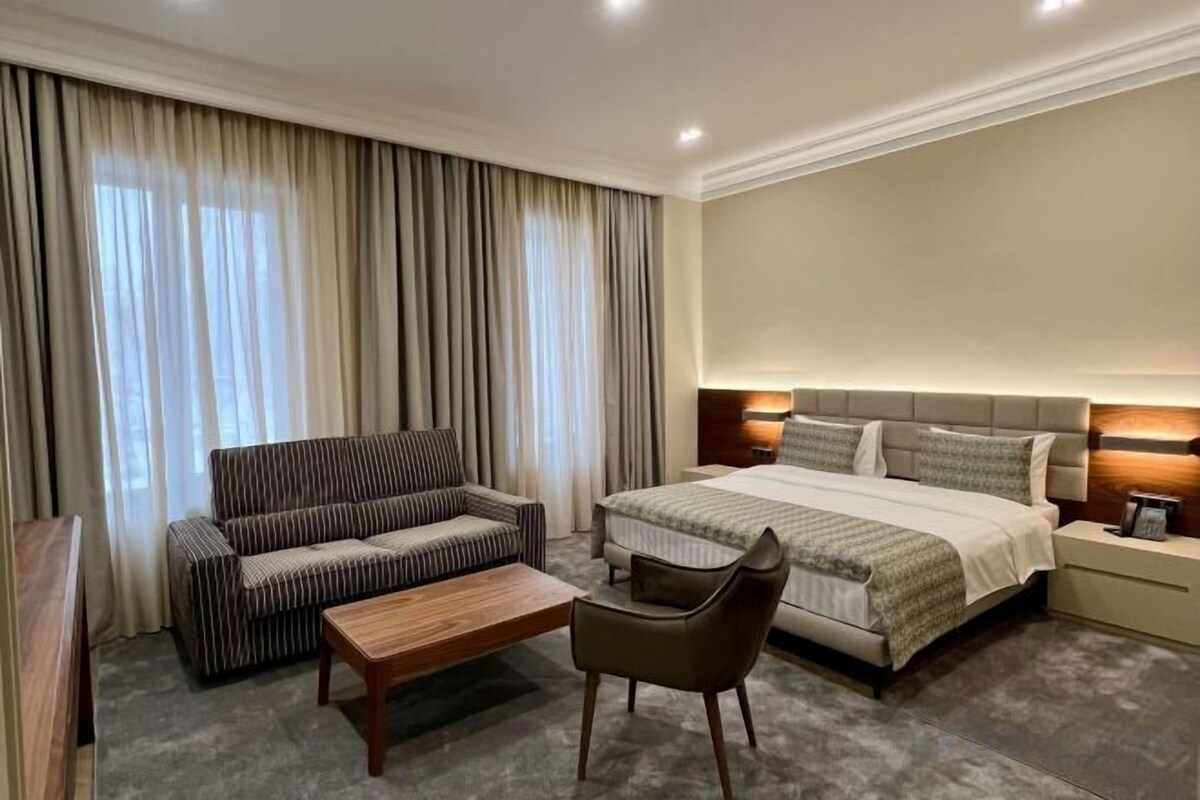 A hotel room with a bed, a sofa, a chair, a small table, and a nightstand with a lamp.