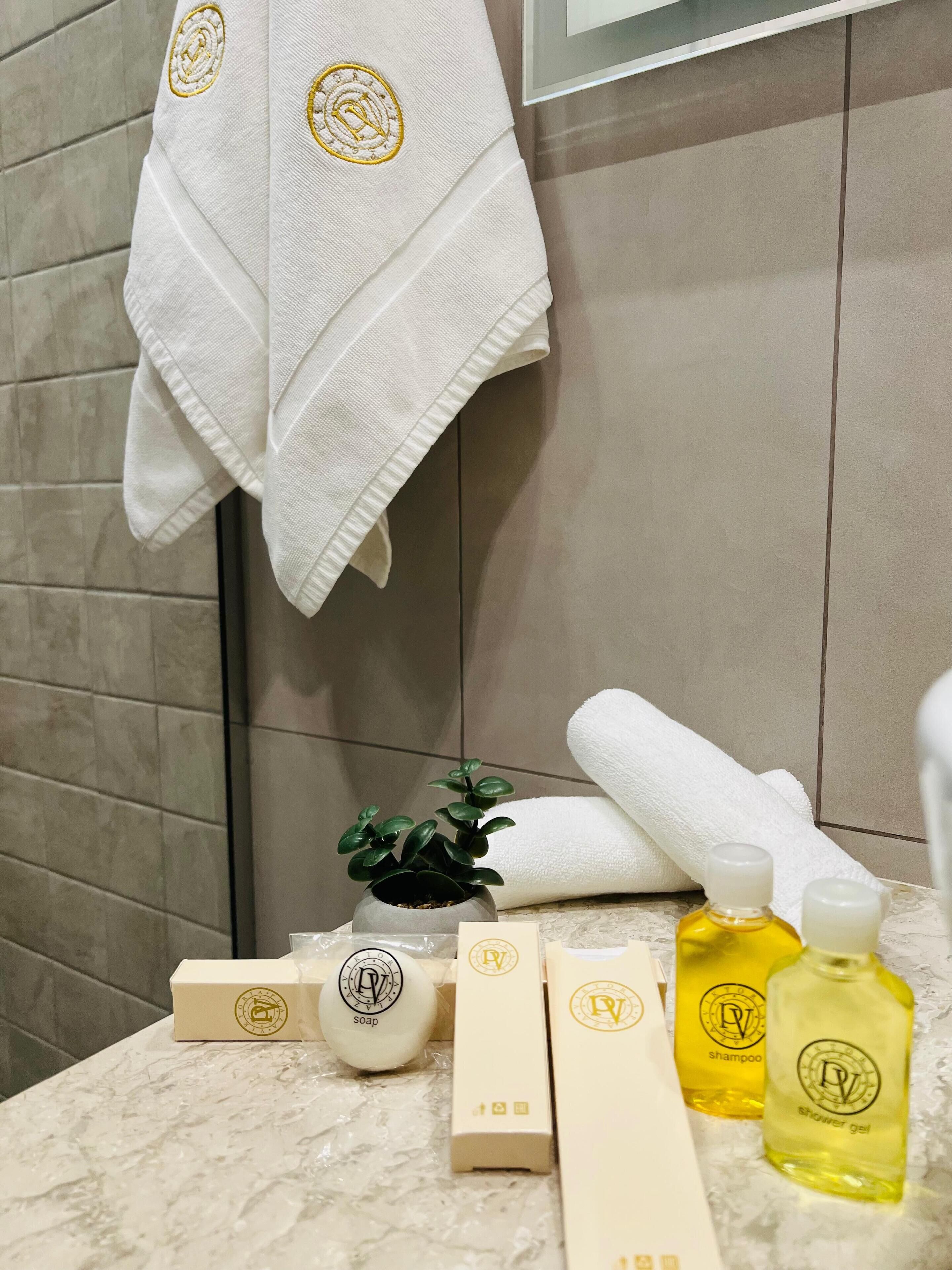 Une salle de bain avec une serviette blanche suspendue à un porte-serviettes, une petite plante en pot et divers articles de toilette, notamment du savon, du shampoing et du gel douche, disposés sur un comptoir en marbre.