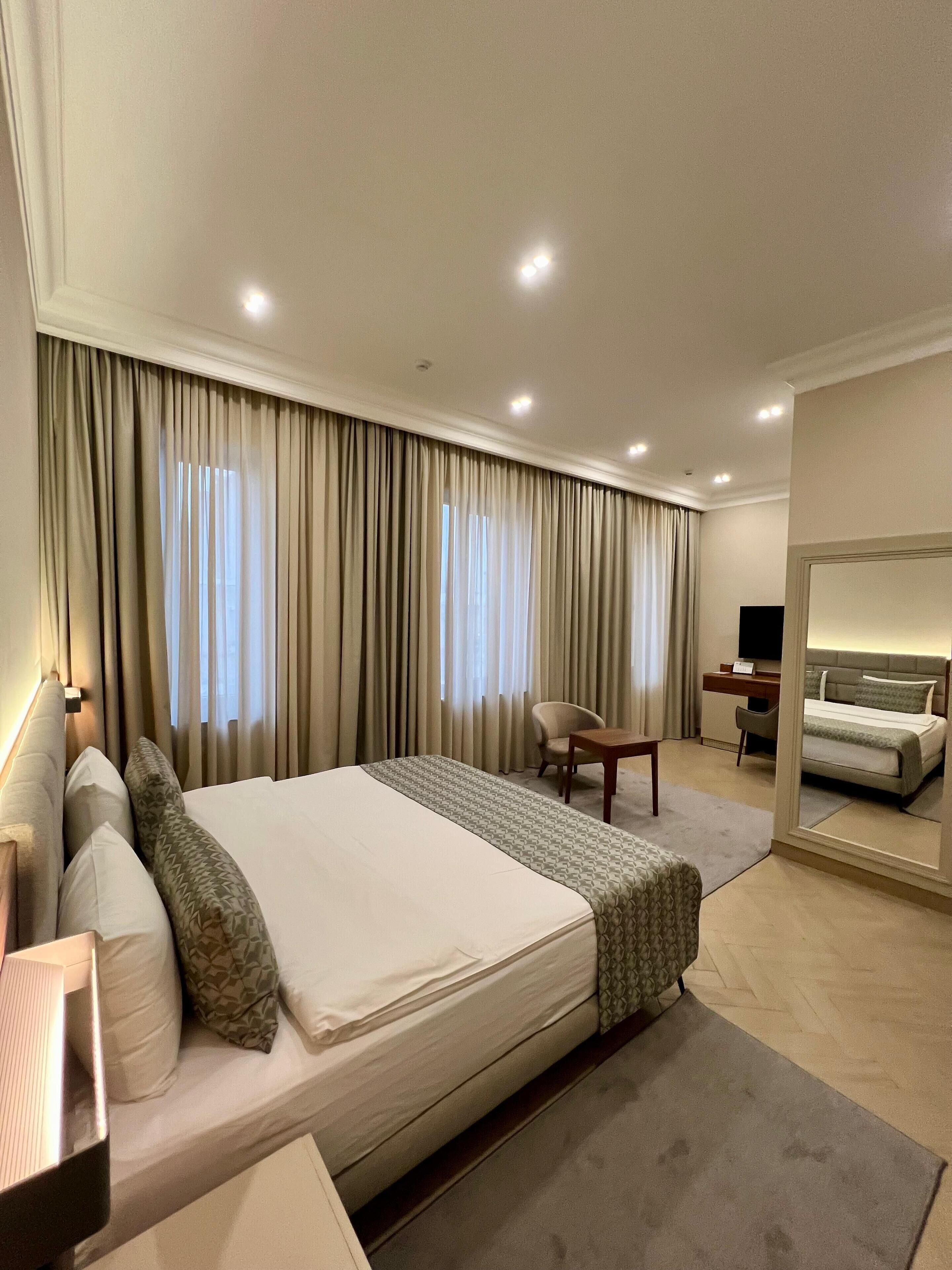 Habitación de hotel con una cama grande, un escritorio, una silla, un espejo y dos ventanas con cortinas.