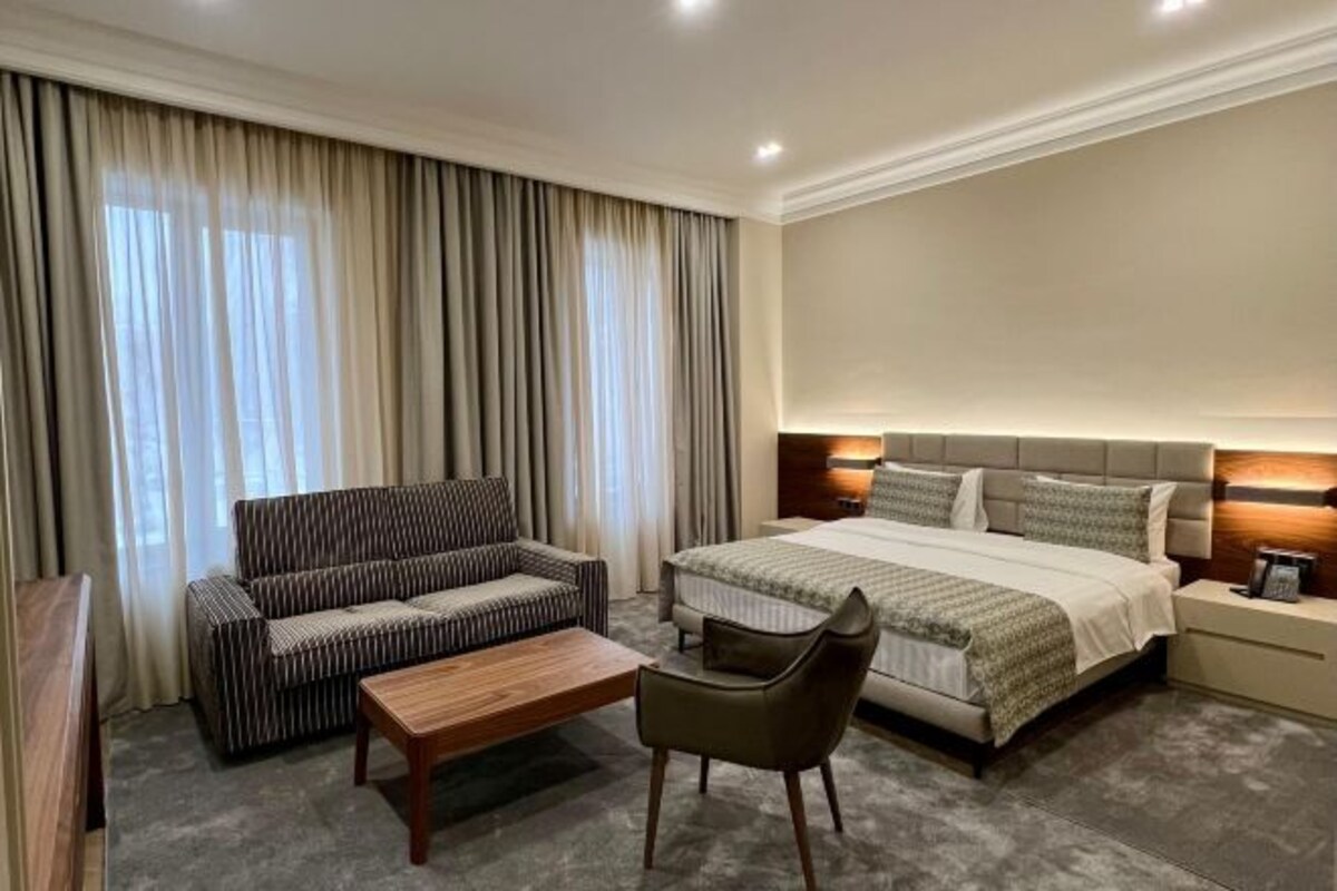 Habitación de hotel con cama, sofá, sillón, mesita de noche y ventana con cortinas.