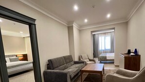 Un vaste espace de vie comprenant un canapé, un fauteuil et une table basse. On aperçoit un lit au fond de la pièce, et un bureau avec une chaise se trouve près d’une fenêtre.