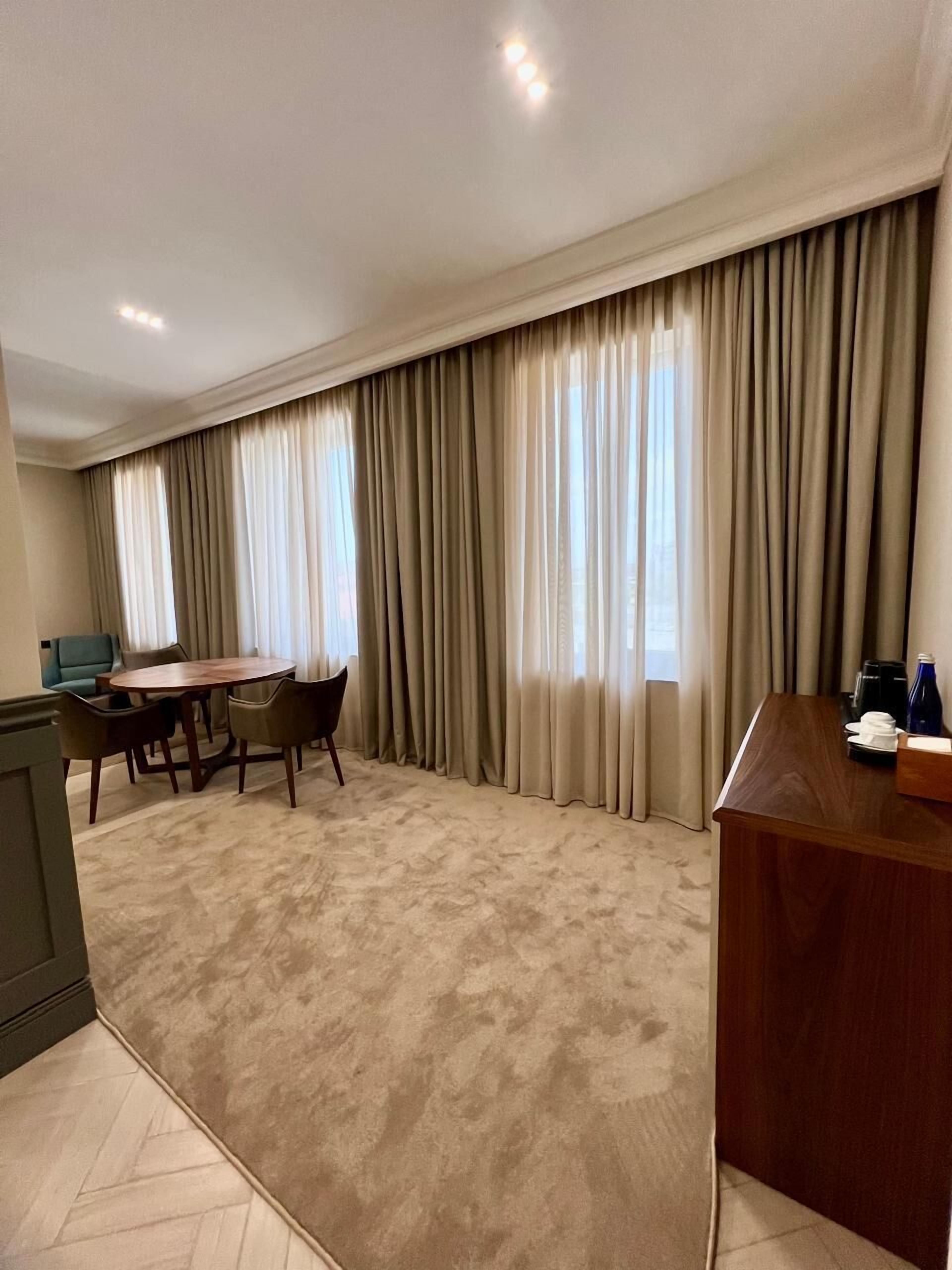 A hotel room with a large window, a dining area with a table and chairs, and a wooden desk with a coffee maker.