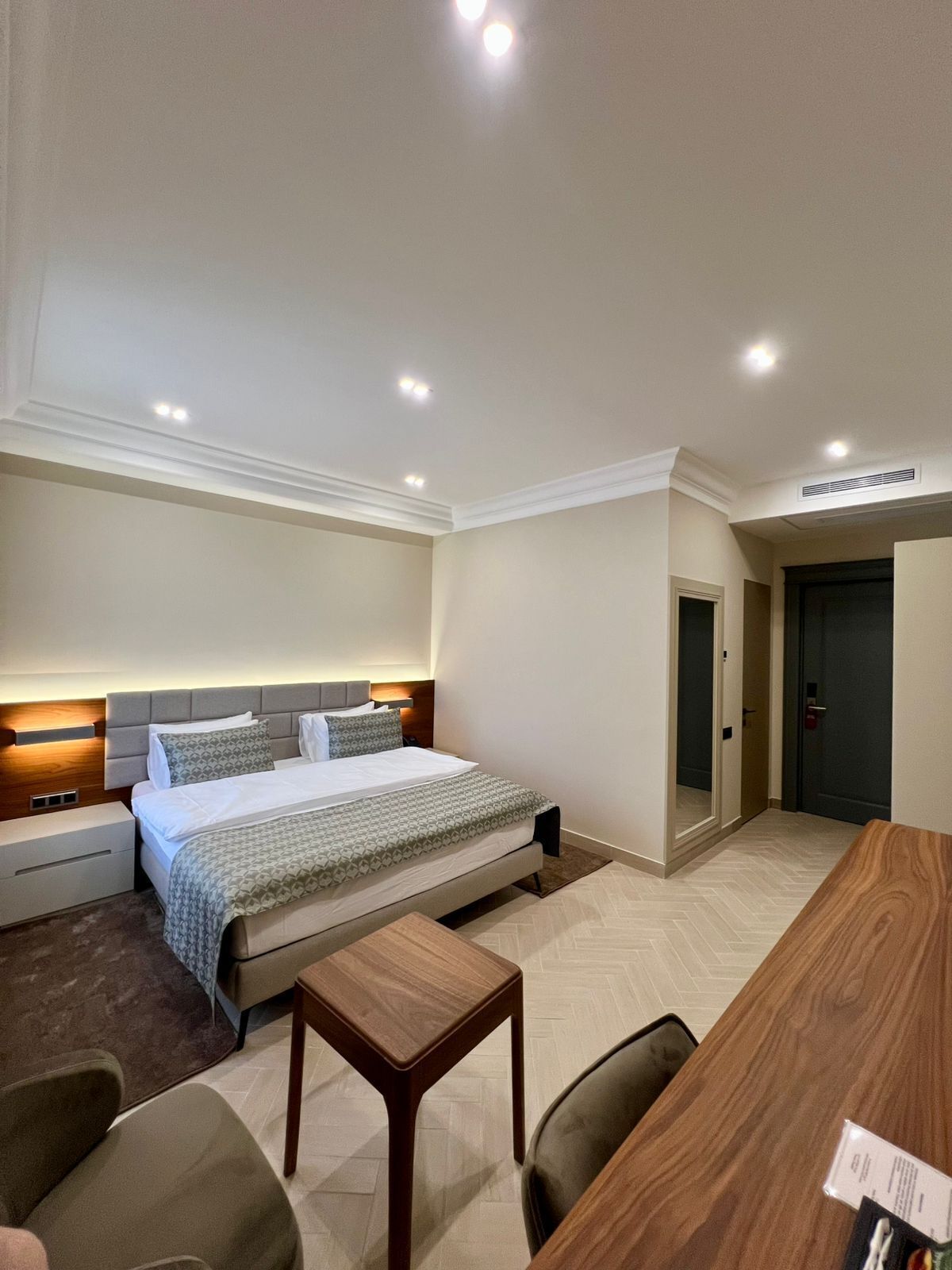 Una habitación de hotel con una cama grande, un escritorio de madera, una silla, una mesita y una puerta.