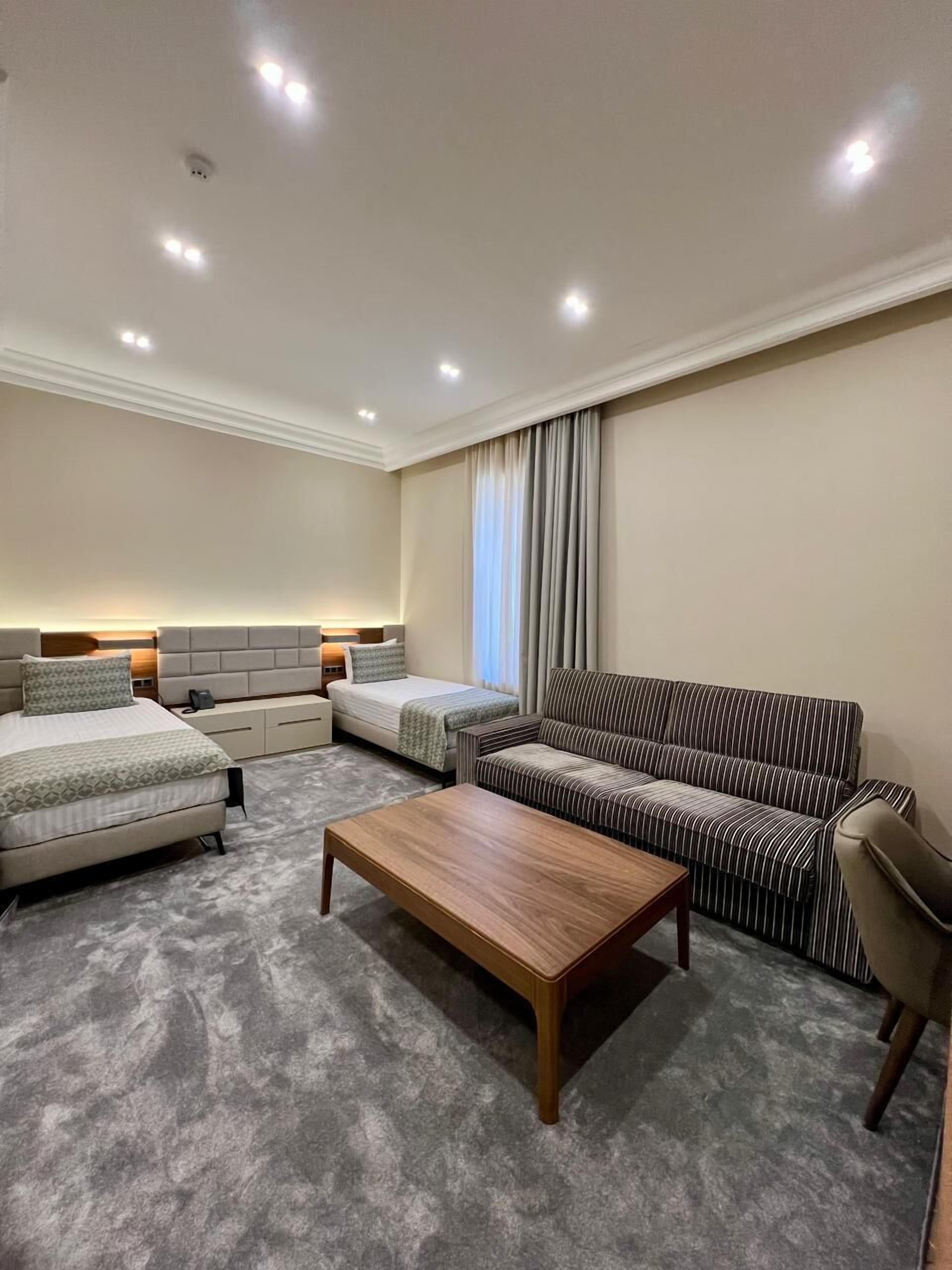 A hotel room with a bed, two bedside tables, a sofa, a coffee table, and a large window with curtains.