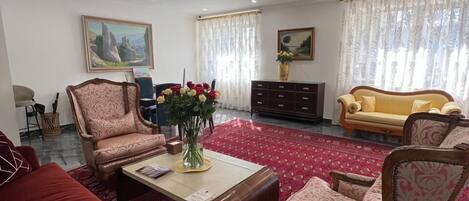 A spacious living room with a red sofa, patterned armchairs, a coffee table, and a yellow sofa. There are two paintings on the walls, a vase with flowers on the coffee table, and a patterned rug on the floor.