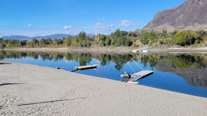 Une rivière paisible où des bateaux sont amarrés sur la rive sablonneuse, entourée d’arbres et avec des montagnes en arrière-plan.
