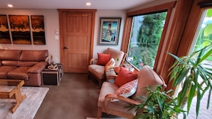 Un salon chaleureux avec un canapé en cuir marron, une table basse en bois et un fauteuil rembourré. Deux tableaux ornent le mur, ainsi qu’une porte en bois et une fenêtre donnant sur un paysage verdoyant.