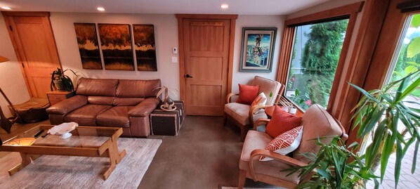 A cozy living room with a brown leather sofa, a wooden coffee table, and a cushioned armchair. There are two paintings on the wall, a wooden door, and a window with a view of greenery.