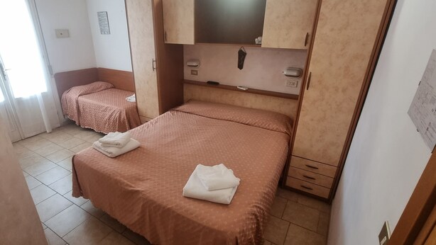 Una stanza con due letti, ognuno con una coperta rosa e un asciugamano bianco piegato. Sopra i letti c'è un armadietto in legno e un tavolino con un telecomando.