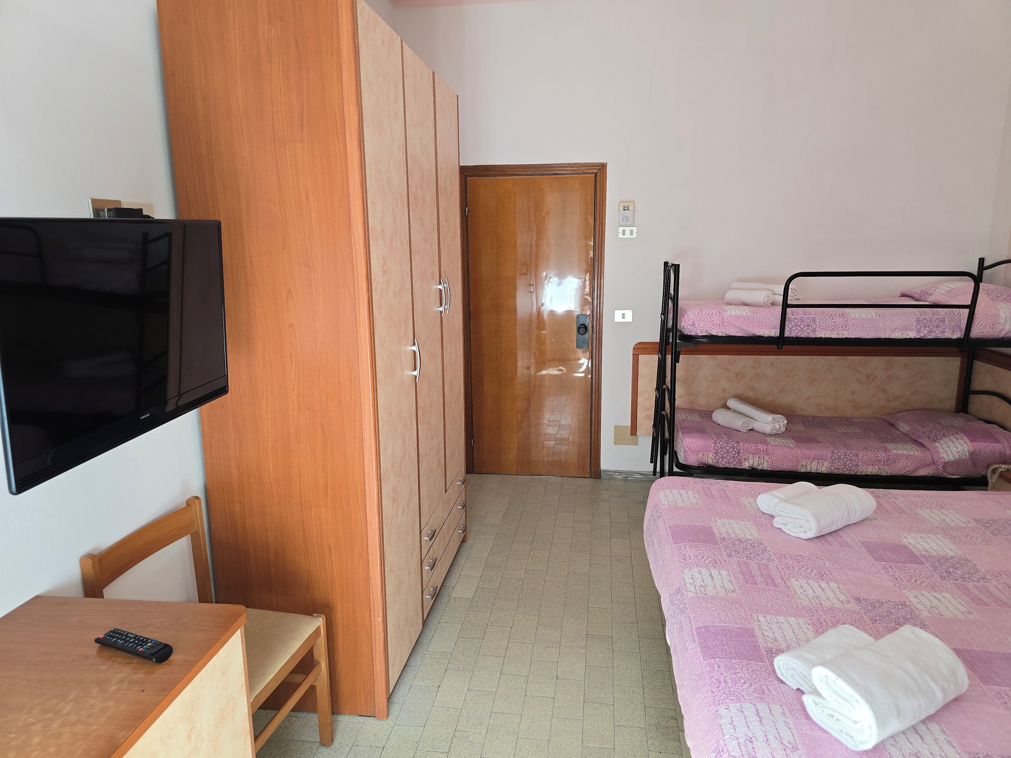 Una habitación con un armario de madera, un escritorio con control remoto, una cama con ropa de cama rosa y una litera con ropa de cama similar.