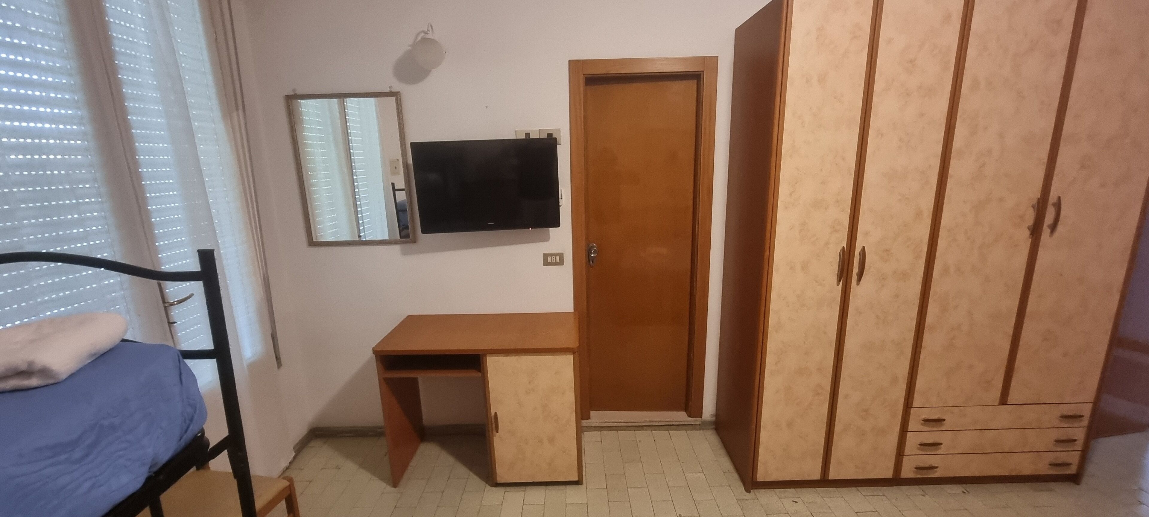 A bedroom with a bed, a wooden desk, a wardrobe, a television, and a mirror.