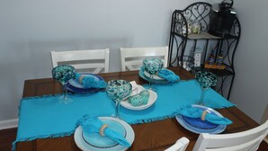 Une table à manger est dressée avec des assiettes, des verres et des serviettes bleues et blanches. Des chaises blanches sont disposées autour de la table.