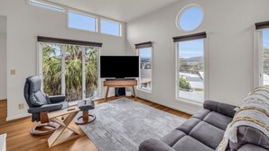 A spacious living room with a large flat-screen TV, comfortable seating, and large windows offering views of palm trees and a cityscape.