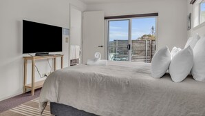 A modern bedroom with a large bed, a flat-screen TV on a wooden console table, and a balcony with a view.