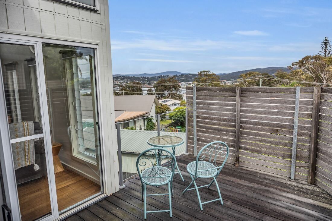 Un balcon doté d’une terrasse en bois, de deux chaises en métal et d’une porte vitrée donnant sur une pièce.