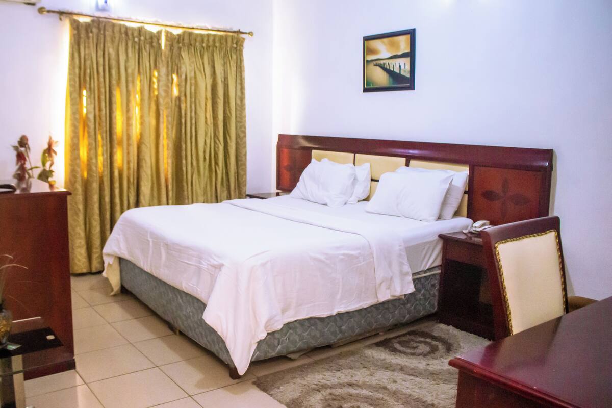 Ein Hotelzimmer mit Bett, Nachttisch, Schreibtisch, Stuhl, Vorhang und einem Bild an der Wand.