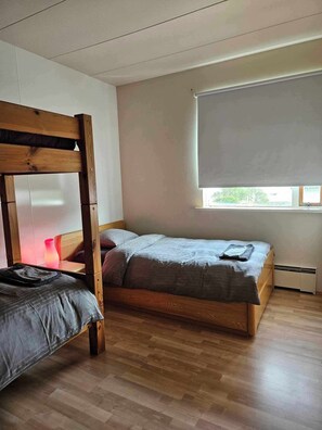 Un dormitorio con una cama litera, una mesita de noche con lámpara, una ventana con persiana y un radiador.
